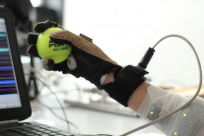 A hand wearing a glove holds a tennis ball. The arm is monitored and information appears on a computer screen.