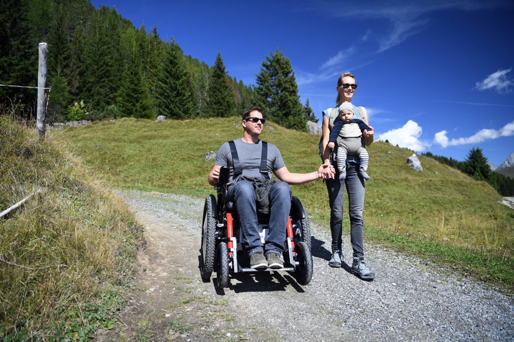 Una coppia sta facendo un'escursione in montagna, l'uomo sulla sedia a rotelle tiene la mano della donna che porta il bambino davanti a sé.