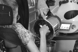 A wheelchair user cooks using a food processor. A third party helps him.